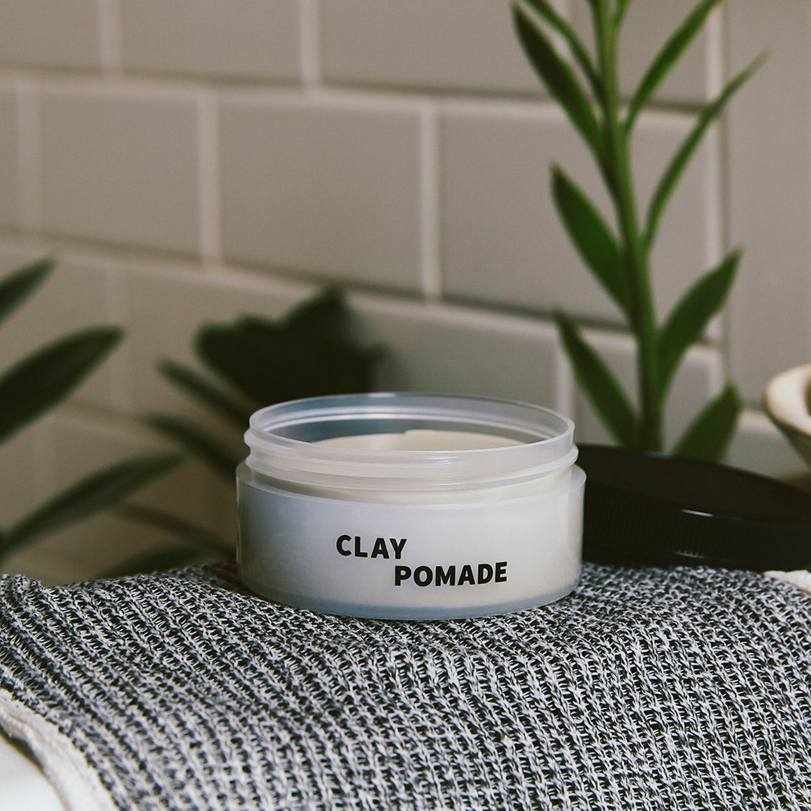 Open jar of Clay Pomade on towel on a sink with plants in the background