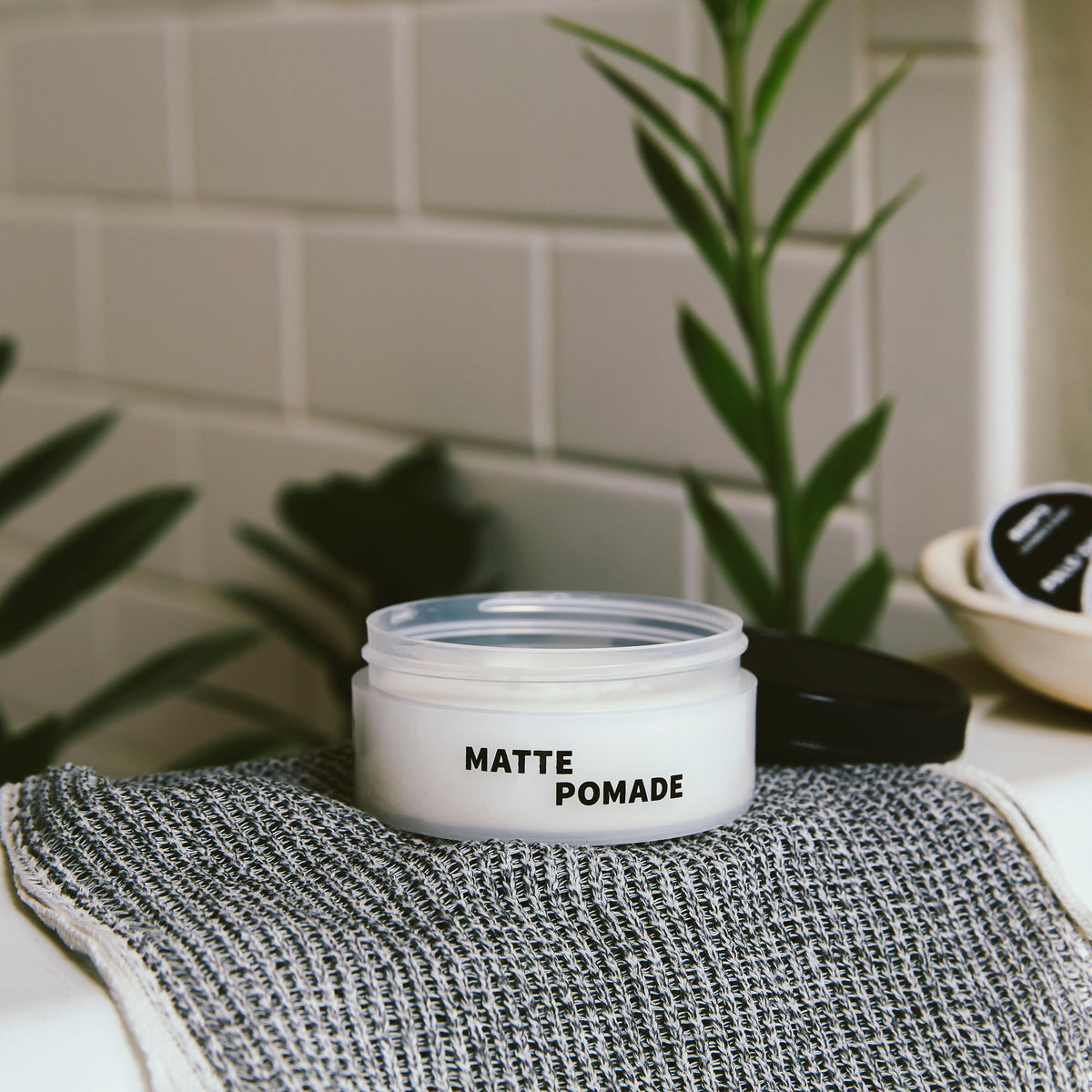 Open jar of Matte Pomade on towel on a sink with plants in the background